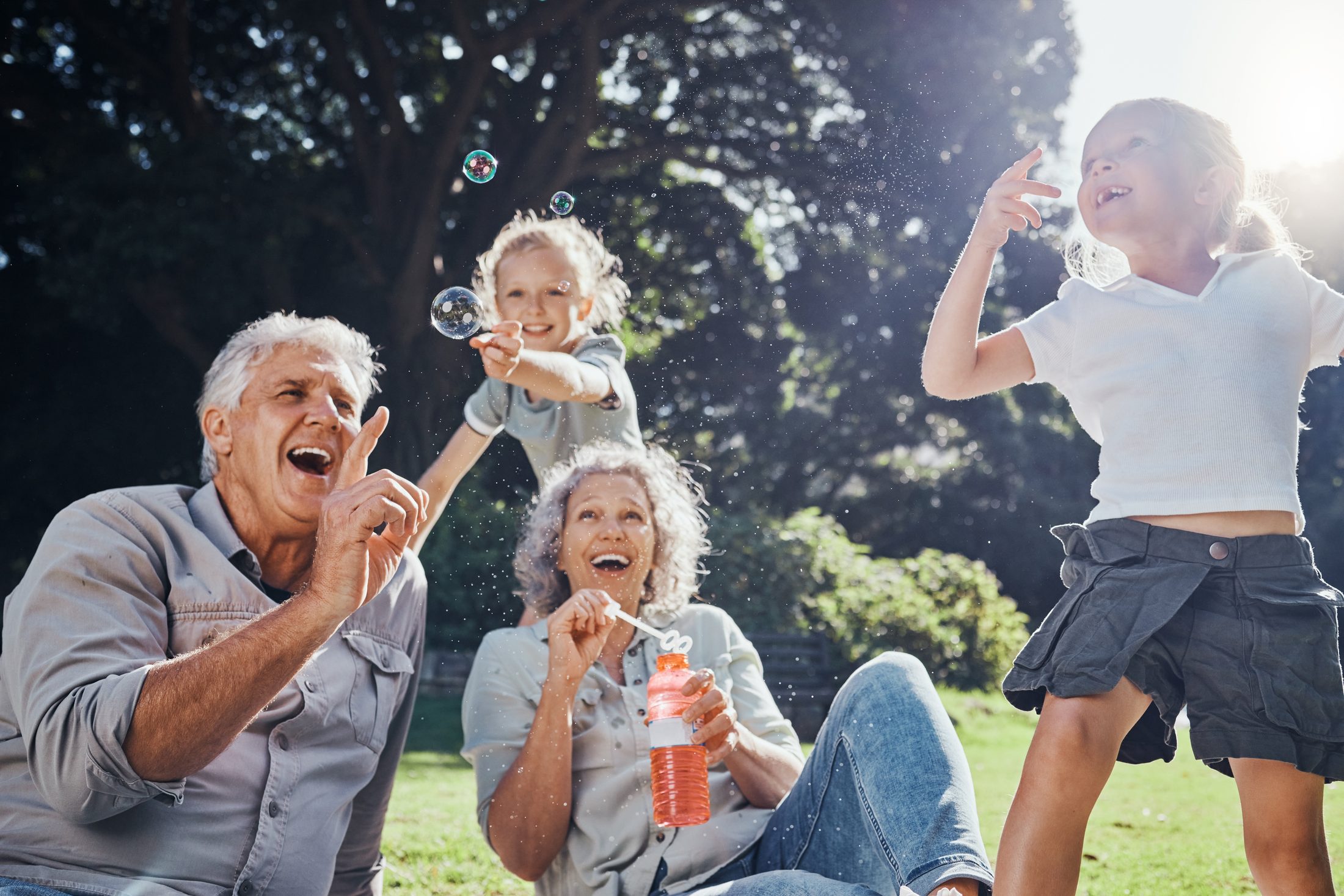 Grandparents laughing with their two grandchildren while blowing bubbles in a sunlit park